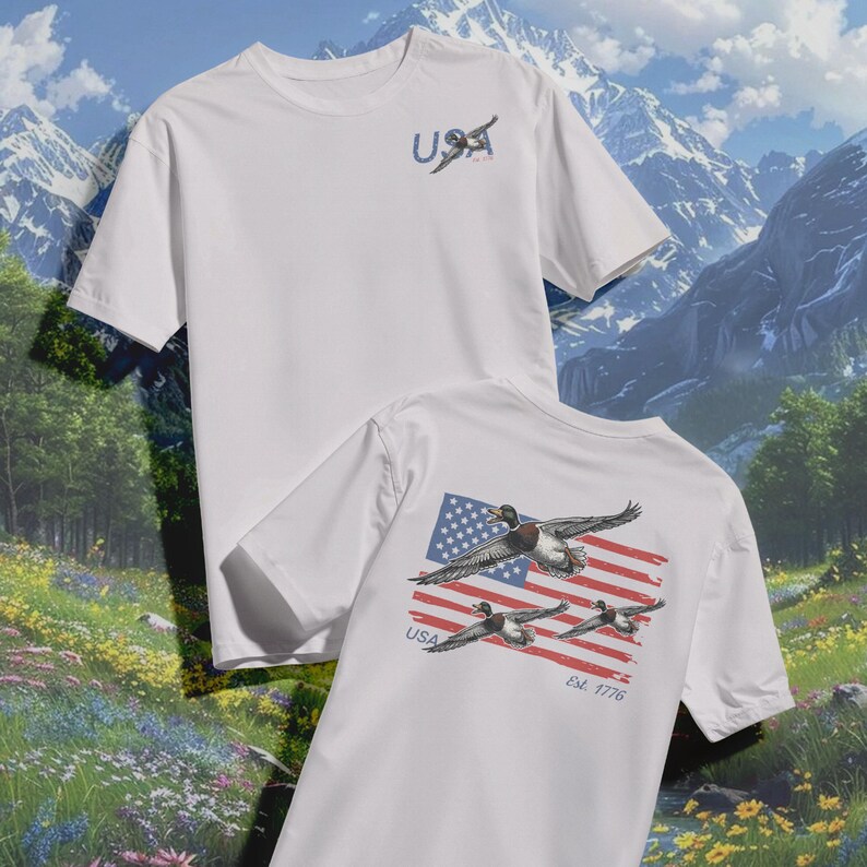 May include: White t-shirts with a patriotic design. One shirt features the word "USA" with a duck graphic on the front. The other shirt displays a duck graphic over an American flag design on the back, with "USA" and "Est. 1776".