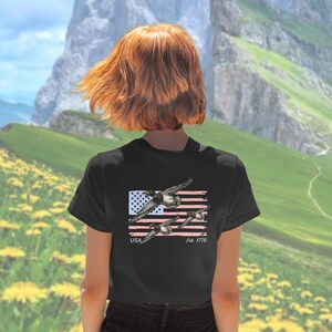 May include: Black t-shirt featuring a graphic of flying ducks over a distressed American flag. The shirt includes the text "USA Est. 1776". The design is in shades of gray, white, and red.