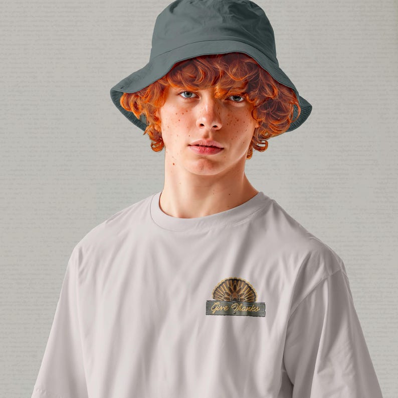 May include: A person wearing a light gray t-shirt and a dark gray bucket hat. The t-shirt features a decorative emblem with the text "Give Thanks". The emblem is a half-circle design with a turkey fan.