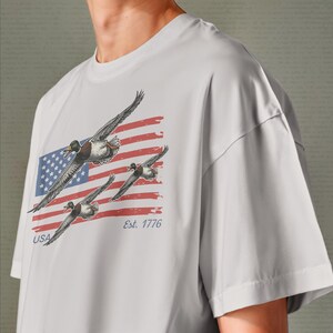 May include: A light gray t-shirt featuring a graphic of three ducks in flight against a backdrop of the American flag. The flag design includes red and white stripes, a blue square with white stars, and the text "USA" and "Est. 1776".