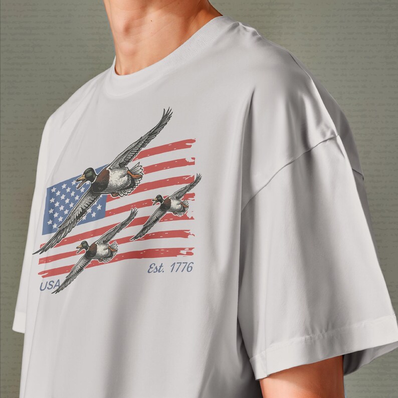 May include: A light gray t-shirt featuring a graphic of three ducks in flight against a backdrop of the American flag. The flag design includes red and white stripes, a blue square with white stars, and the text "USA" and "Est. 1776".