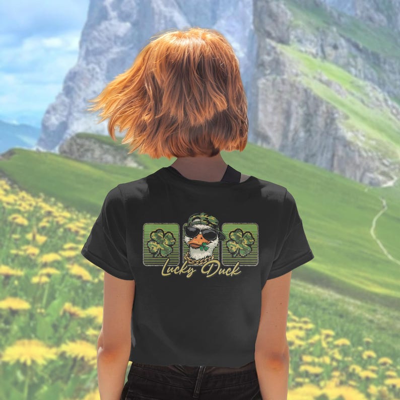 May include: Black t-shirt featuring a graphic of a duck wearing sunglasses and a camouflage hat, with a gold chain and a shamrock. The text "Lucky Duck" is below the graphic, with shamrock designs on either side.