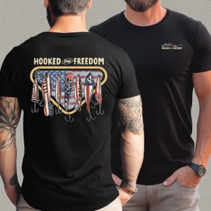 May include: Black t-shirt with a graphic on the back featuring fishing lures with an American flag design and the text "HOOKED ON FREEDOM". A smaller version of the graphic is on the front.