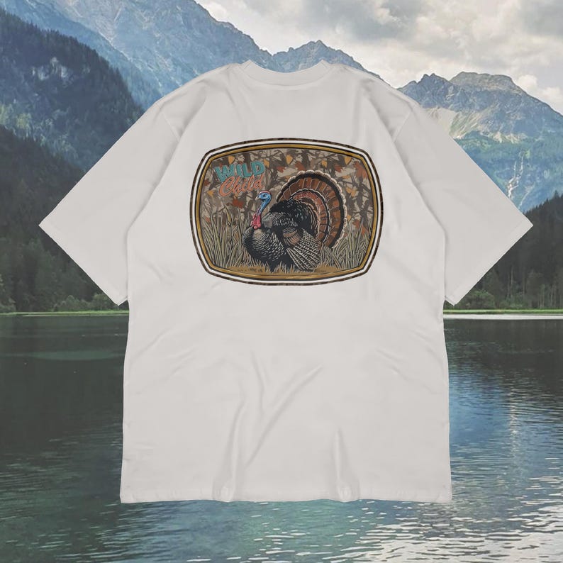 May include: White t-shirt featuring a detailed illustration of a turkey with its feathers fanned out. The graphic is set within a brown and tan oval frame, with the words "Wild Game" above the turkey. The shirt has short sleeves and a crew neck.