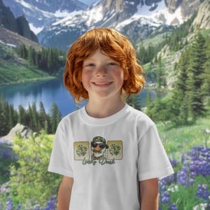 May include: White t-shirt featuring a cartoon duck wearing sunglasses, a camouflage hat, and a gold chain. The design includes shamrocks and the text "Lucky Duck". The background shows a scenic mountain landscape with a lake.
