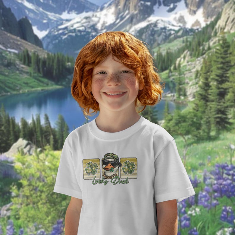 May include: White t-shirt featuring a cartoon duck wearing sunglasses, a camouflage hat, and a gold chain. The design includes shamrocks and the text "Lucky Duck". The background shows a scenic mountain landscape with a lake.