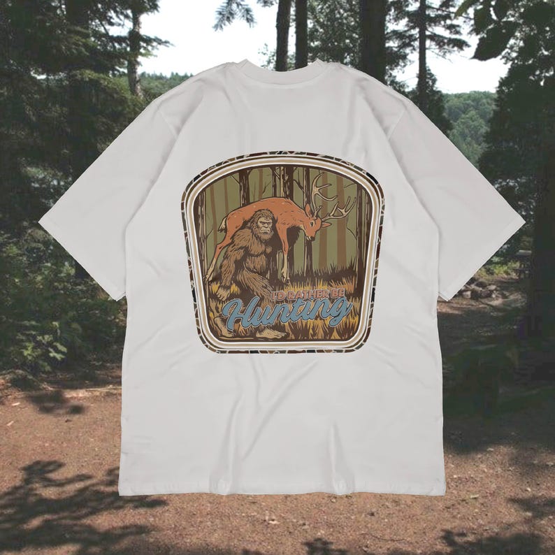 May include: White t-shirt featuring a graphic of Bigfoot and a deer in a forest scene. The design includes the text "To Dare to Hunting" in a vintage style. The shirt is made of a soft, comfortable material.