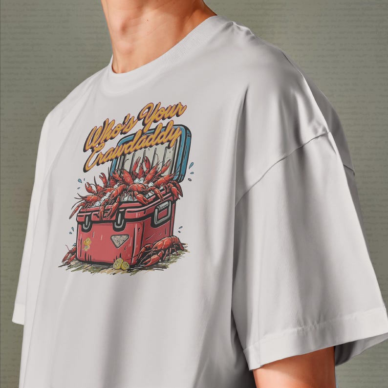 以下が含まれることがあります： 白いTシャツ。赤いクーラーボックスからザリガニがあふれ出ているグラフィック。クーラーボックスには青い蓋があり、テキスト「Who's Your Crawdaddy」は黄色の様式化されたフォント。デザインにはレモンと水滴が含まれています。