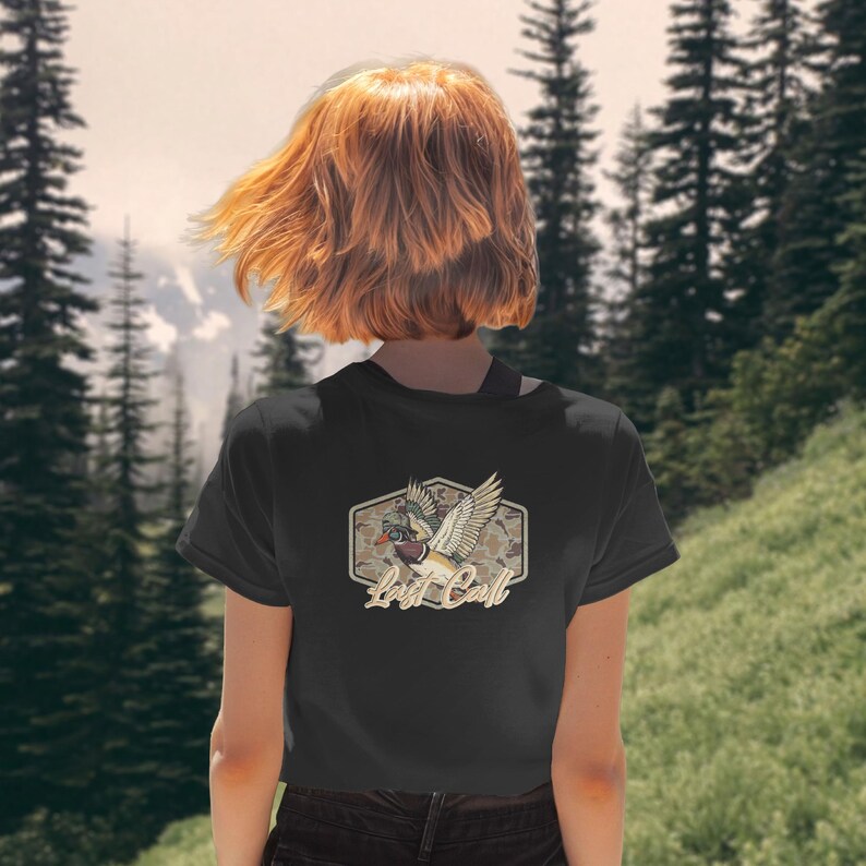 May include: Black t-shirt featuring a duck graphic with the text "Lost Call" in a vintage style. The design includes a duck in flight with outstretched wings, set against a camouflage pattern. The shirt is a classic crew neck.