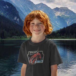 Pu&ograve; includere: Una maglietta nera con una grafica di un gambero rosso su un amo da pesca, incorniciata con le parole "Crawfish Season". La maglietta &egrave; indossata da una persona con i capelli rossi, sullo sfondo di montagne e un lago.