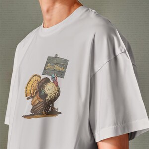 May include: A light gray t-shirt featuring a colorful illustration of a turkey holding a wooden sign that reads "Give Thanks". The turkey has brown, tan, and orange feathers. The shirt is a casual style.