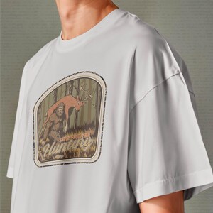 May include: Light gray t-shirt featuring a forest scene with a deer and Bigfoot, with the text "I'd Rather Be Hunting" in a brown and tan design. The shirt has short sleeves and a crew neck.