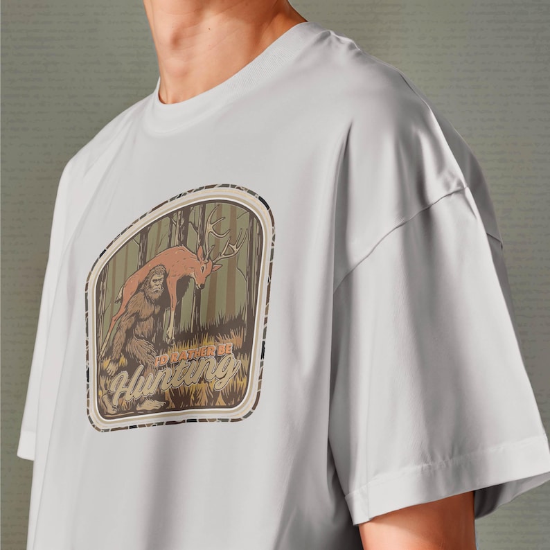 May include: Light gray t-shirt featuring a forest scene with a deer and Bigfoot, with the text "I'd Rather Be Hunting" in a brown and tan design. The shirt has short sleeves and a crew neck.