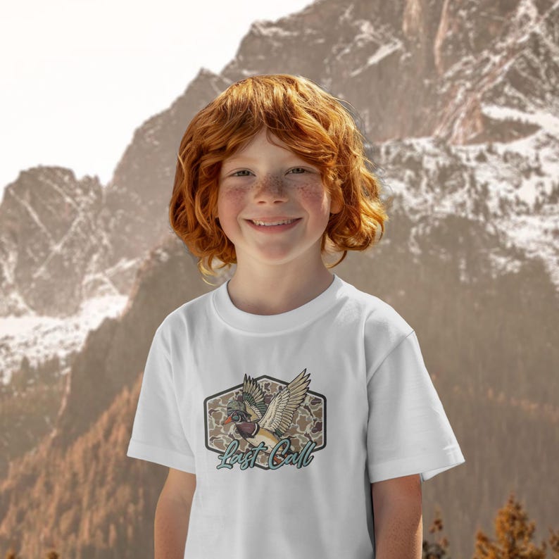 May include: White t-shirt featuring a graphic of a duck with outstretched wings, a camouflage background, and the text "Last Call" in a teal script. The shirt is worn by a person with red hair and freckles, smiling in front of a mountain backdrop.