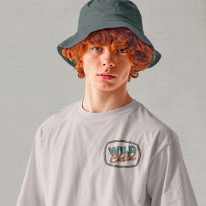 May include: A light gray t-shirt with a retro-style graphic that reads "Wild Child" in a rectangular design. The person in the image is wearing a gray bucket hat.