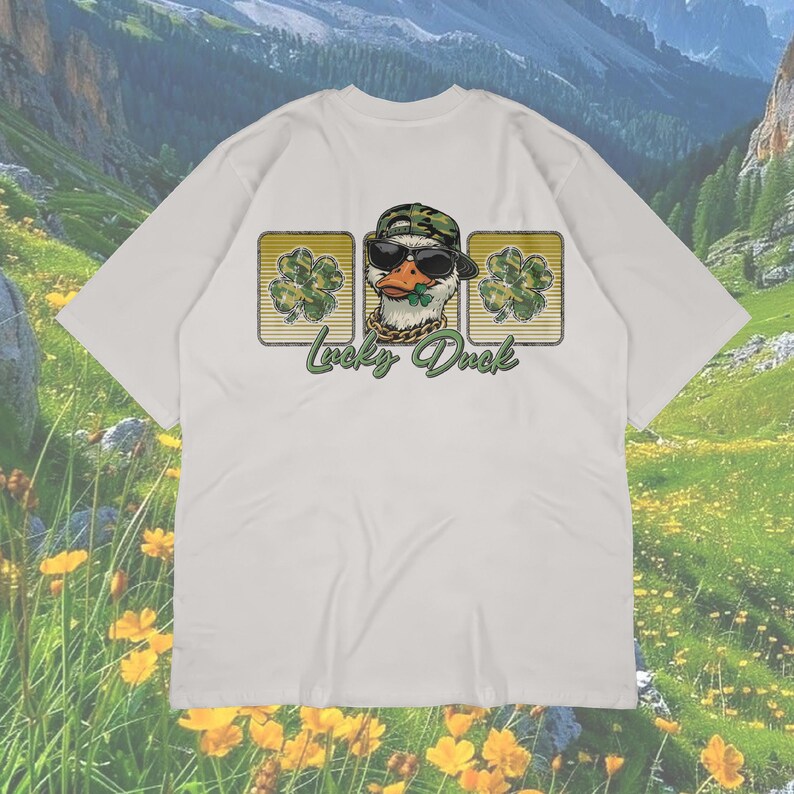 May include: White t-shirt featuring a graphic of a duck wearing sunglasses and a camouflage hat, with a gold chain and a shamrock. The text "Lucky Duck" is below the duck image, with two shamrock designs on either side.