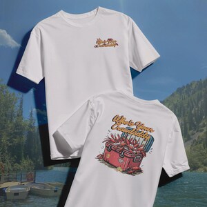 以下が含まれることがあります： 2つの白いTシャツ。1つのシャツには、前面と背面に「Who's Your Crawdaddy?」というテキストがあります。シャツの背面には、ザリガニで溢れる赤いクーラーボックスが描かれています。前面には同じテキストが小さなフォントで表示されています。