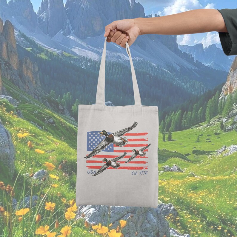 May include: A white tote bag featuring a graphic of flying ducks in front of a stylized American flag. The flag has red and white stripes with a blue field of stars. The text "USA" and "Est. 1776" are printed below the flag.
