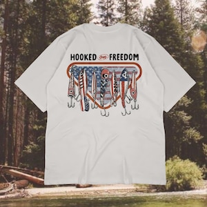 May include: White t-shirt featuring a graphic of fishing lures in red, white, and blue, with the American flag design. The text "HOOKED ON FREEDOM" is above the lures. A casual wear item.