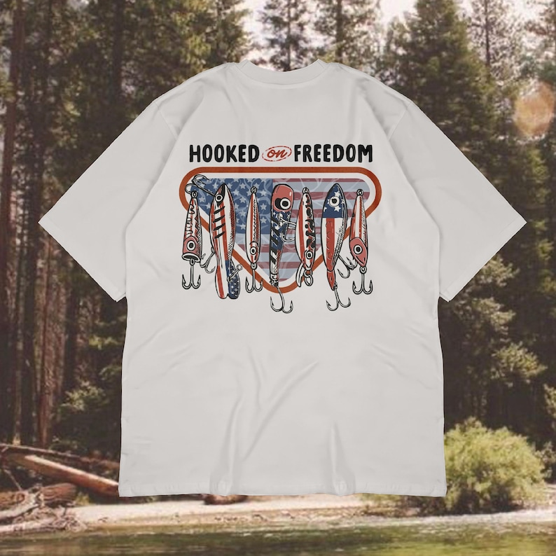 May include: White t-shirt featuring a graphic of fishing lures in red, white, and blue, with the American flag design. The text "HOOKED ON FREEDOM" is above the lures. A casual wear item.