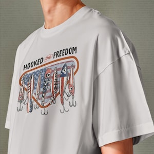 May include: A pale grey t-shirt with a graphic of fishing lures in red, white, and blue. The lures are set against a backdrop of the American flag. The text "HOOKED ON FREEDOM" is printed above the lures.