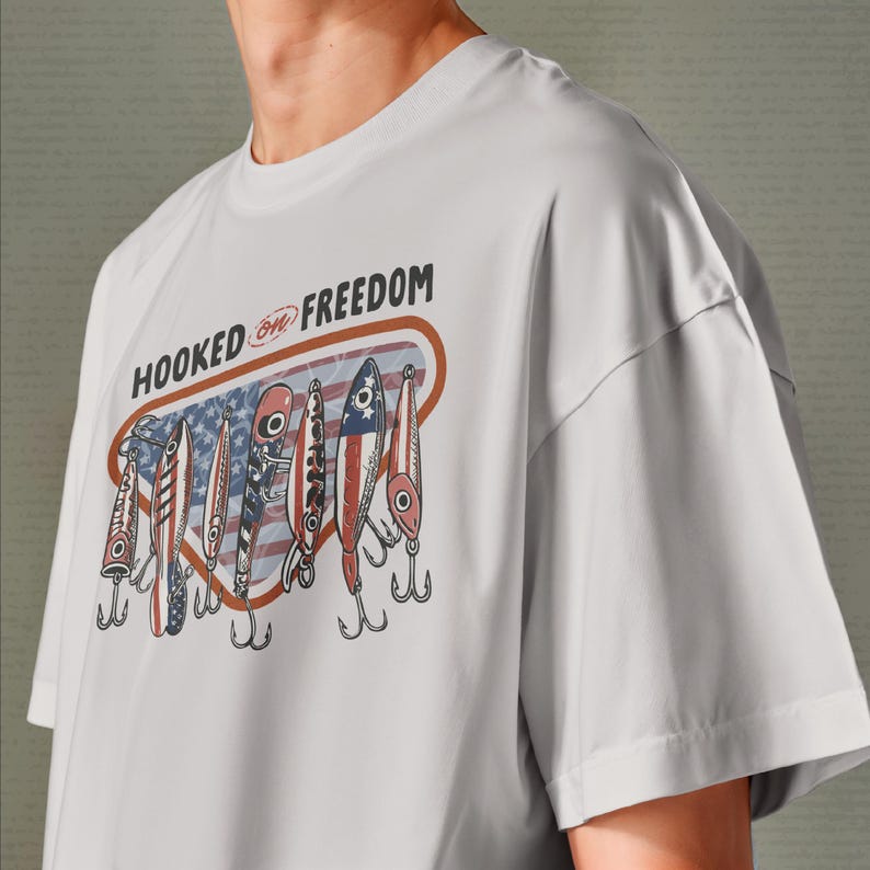 May include: A pale grey t-shirt with a graphic of fishing lures in red, white, and blue. The lures are set against a backdrop of the American flag. The text "HOOKED ON FREEDOM" is printed above the lures.