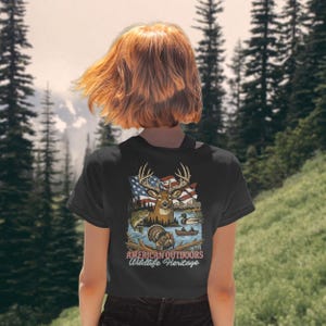 以下が含まれることがあります： 黒いTシャツ。鹿、アメリカ国旗、魚、アヒル、七面鳥のグラフィック。グラフィックの下には「AMERICAN OUTDOORS Wildlife Heritage」の文字。