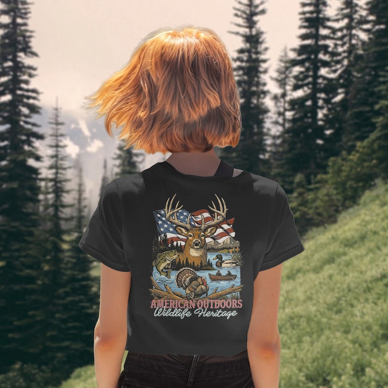 以下が含まれることがあります： 黒いTシャツ。鹿、アメリカ国旗、魚、アヒル、七面鳥のグラフィック。グラフィックの下には「AMERICAN OUTDOORS Wildlife Heritage」の文字。