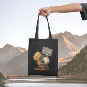 May include: Black tote bag featuring a colorful illustration of a turkey holding a wooden sign that reads "Give Thanks." The turkey has a vibrant display of feathers in shades of yellow, orange, and brown. The bag is held up against a backdrop of mountains and water.