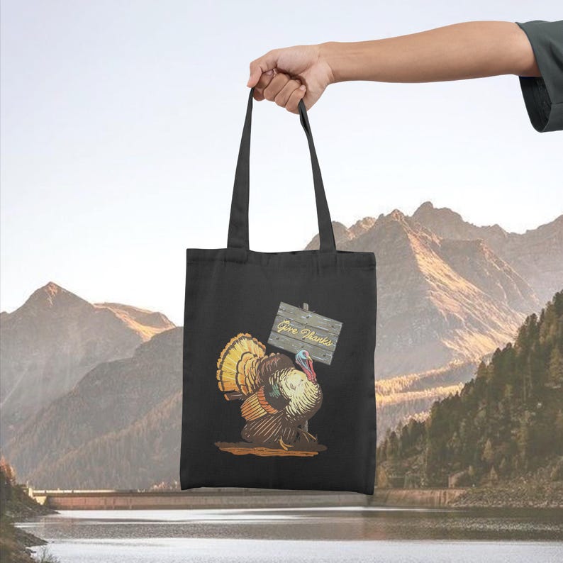 May include: Black tote bag featuring a colorful illustration of a turkey holding a wooden sign that reads "Give Thanks." The turkey has a vibrant display of feathers in shades of yellow, orange, and brown. The bag is held up against a backdrop of mountains and water.