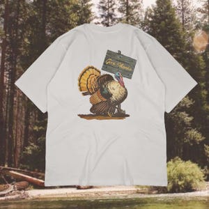 May include: White t-shirt featuring a colorful illustration of a turkey holding a wooden sign that reads "Give Thanks." The turkey has brown, orange, and yellow feathers. The shirt is a casual wear item.