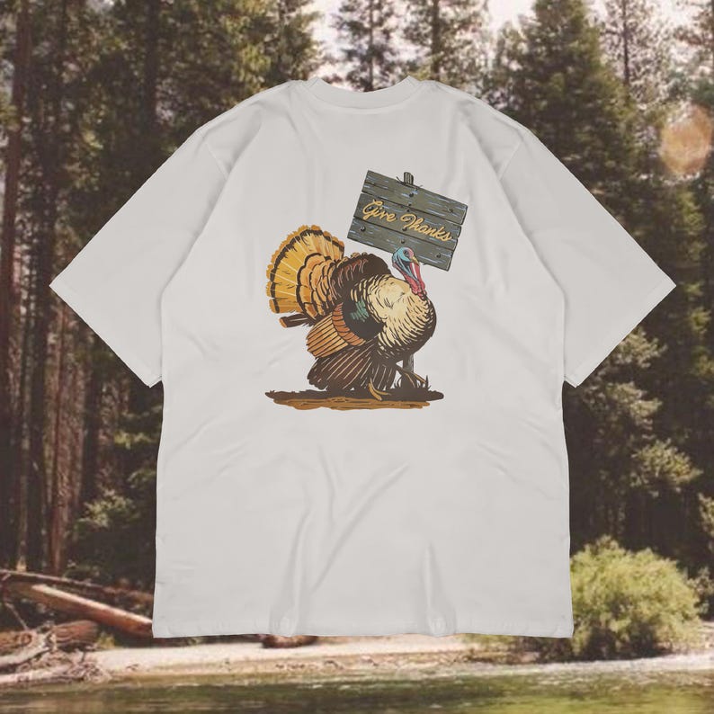 May include: White t-shirt featuring a colorful illustration of a turkey holding a wooden sign that reads "Give Thanks." The turkey has brown, orange, and yellow feathers. The shirt is a casual wear item.