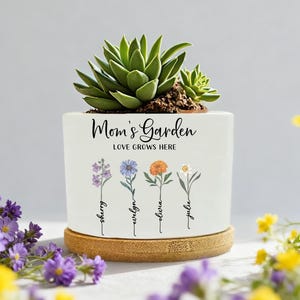 Puede incluir: Una maceta de cerámica blanca con el texto "Mom's Garden LOVE GROWS HERE" e ilustraciones florales. La maceta contiene una planta suculenta verde y se apoya en un posavasos redondo de bambú. Los nombres "Sherry, Evelyn, Olivia, Julie" están debajo.