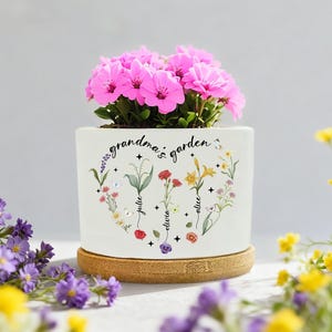 May include: A white ceramic plant pot with the words "grandma's garden" and floral illustrations. The pot contains bright pink flowers and rests on a round, light brown bamboo coaster. The names "Julie", "Olivia", and "Alice" are written on the pot.