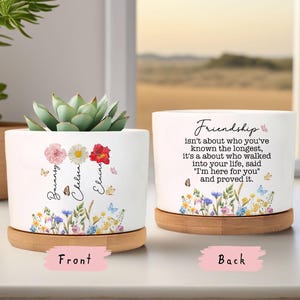 Puede incluir: Maceta de cerámica blanca con una suculenta, con diseños florales y mariposas. La parte delantera muestra nombres y flores, la trasera una cita sobre la amistad. Incluye un platillo de bambú.