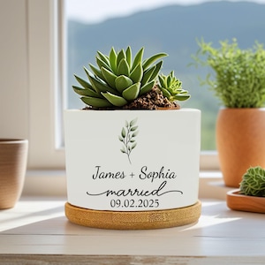 Puede incluir: Una maceta de cerámica blanca con una planta suculenta, sobre un posavasos de bambú. La maceta tiene el texto "James + Sophia married 09.02.2025" y un diseño de hojas. Una pequeña planta verde es visible en el fondo.