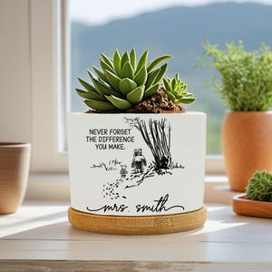 May include: White ceramic planter with a succulent plant, featuring a black illustration of a bear and a smaller animal walking on a path. The text reads "NEVER FORGET THE DIFFERENCE YOU MAKE" and "mrs. smith". The planter sits on a bamboo saucer.