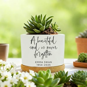 Puede incluir: Una maceta de cerámica blanca con una planta suculenta. La maceta tiene el texto "A beautiful soul is never forgotten" y el nombre "EMMA SWAN 1954-2025". La maceta se asienta sobre una base de madera, con otras plantas en el fondo.