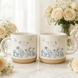 Puede incluir: Dos tazas de cerámica blanquecina con un diseño floral azul. Cada taza tiene un asa y una base marrón claro. Las tazas están decoradas con mariposas, iniciales, un corazón y la fecha 27.01.2026. Rosas blancas rodean las tazas.