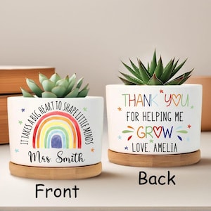 May include: Two white ceramic plant pots with succulents, each on a bamboo saucer. One pot has a rainbow design and the text "It takes a big heart to shape little minds" and "Mrs. Smith". The other says "Thank you for helping me grow, love Amelia".