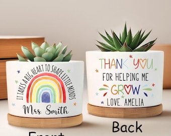 Może przedstawiać: Dwa białe ceramiczne doniczki z sukulentami, każda na bambusowej podstawce. Jedna doniczka ma tęczowy wzór i napis "It takes a big heart to shape little minds" i "Mrs. Smith". Druga mówi "Thank you for helping me grow, love Amelia".
