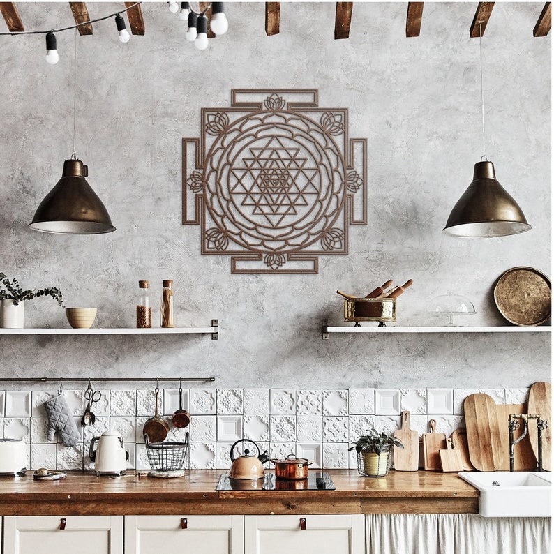 May include: A brown wooden wall hanging with a geometric design, featuring a mandala with a central pyramid shape. The wall hanging is displayed in a kitchen setting with white tile backsplash and wooden countertops.