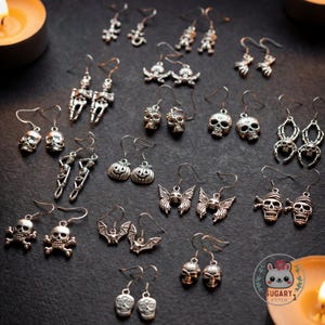 Puede incluir: Una colección de pendientes plateados con temática de Halloween, que incluyen diseños de calaveras, esqueletos, murciélagos, arañas y calabazas. Los pendientes se exhiben sobre una superficie oscura, con algunas velas encendidas en el fondo.