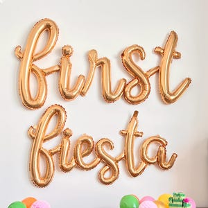 Pode incluir: Balões dourados que formam a frase "first fiesta" numa parede branca. Abaixo, balões coloridos e decorações, incluindo uma pinhata de cacto, estão dispostos sobre uma superfície cinzenta, criando uma cena festiva.