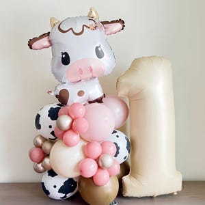 Pode incluir: Um buquê de balões com um balão em forma de vaca, um balão grande com o número um e balões menores em rosa, marrom e branco com padrões de vaca.