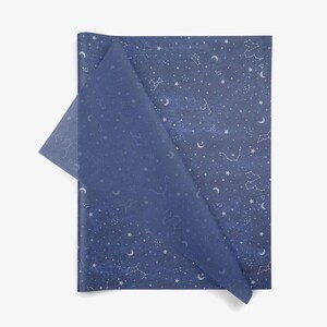Puede incluir: Papel de regalo azul oscuro con un dise&ntilde;o celestial. El papel presenta constelaciones, estrellas y lunas crecientes blancas. Una esquina del papel est&aacute; doblada, revelando una capa transl&uacute;cida con el mismo patr&oacute;n.