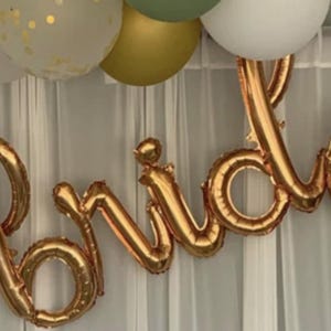 Könnte beinhalten: Ein roségoldener Folienballon-Schriftzug, der das Wort bride in Schreibschrift bildet. Der Ballon ist vor einem weißen, halbtransparenten Vorhang platziert, wobei oben eine Ansammlung von weißen, goldenen und olivgrünen Ballons zu sehen ist.