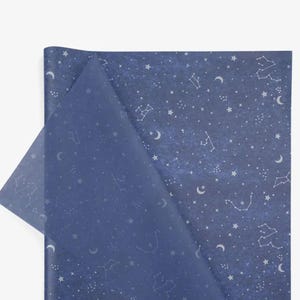 Puede incluir: Papel de regalo azul oscuro con un dise&ntilde;o celestial. Presenta constelaciones, estrellas y lunas crecientes blancas. Una esquina est&aacute; doblada, revelando una capa transl&uacute;cida azul m&aacute;s clara.