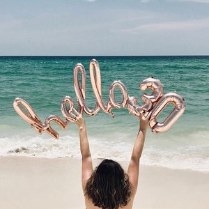 May include: A person holding up a rose gold foil balloon that says "Hello 30" in front of a beach with blue water and white sand.