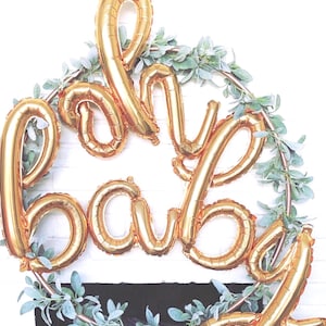 Pode incluir: Balões dourados em forma de letras formam a palavra "baby" numa grinalda circular com vegetação. O conjunto está sobre uma parede de tijolos brancos, ideal para um chá de bebé ou a decoração de um quarto infantil. O ambiente é festivo.